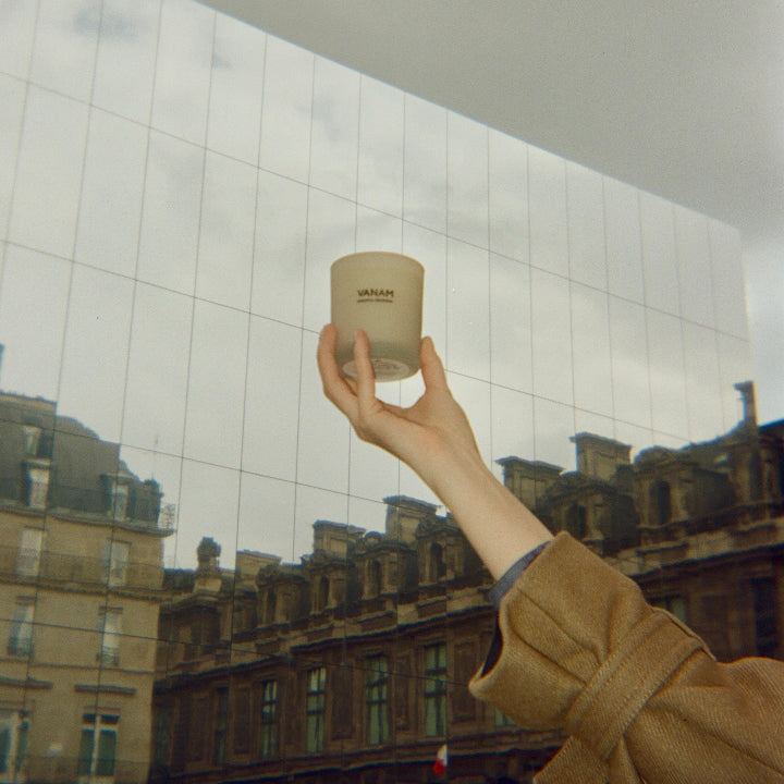Woman's hand holding Vanam beeswax candle in front of a reflective building.